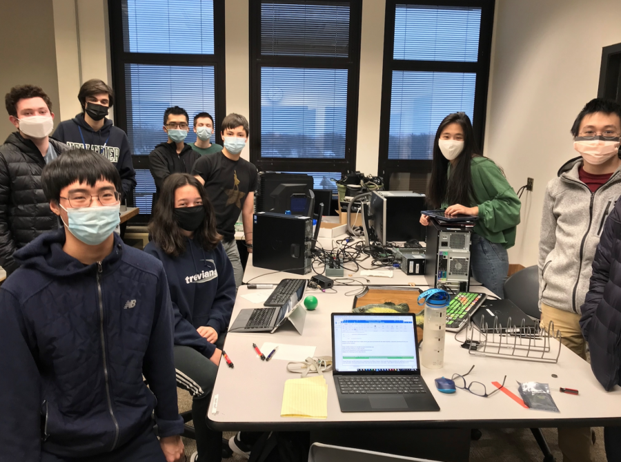 Members of the BinaryHeart Club stand around their workstation filled with computers, monitors, tablets, cords, and wires.
